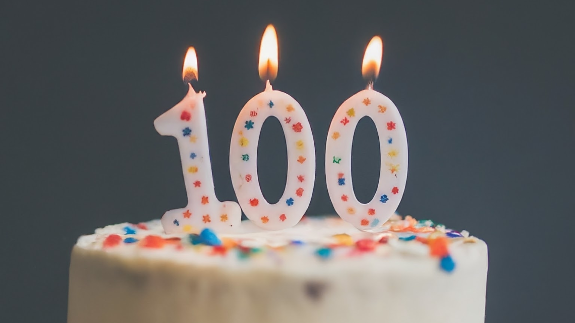 A 100th birthday cake with lit birthday candles that say100